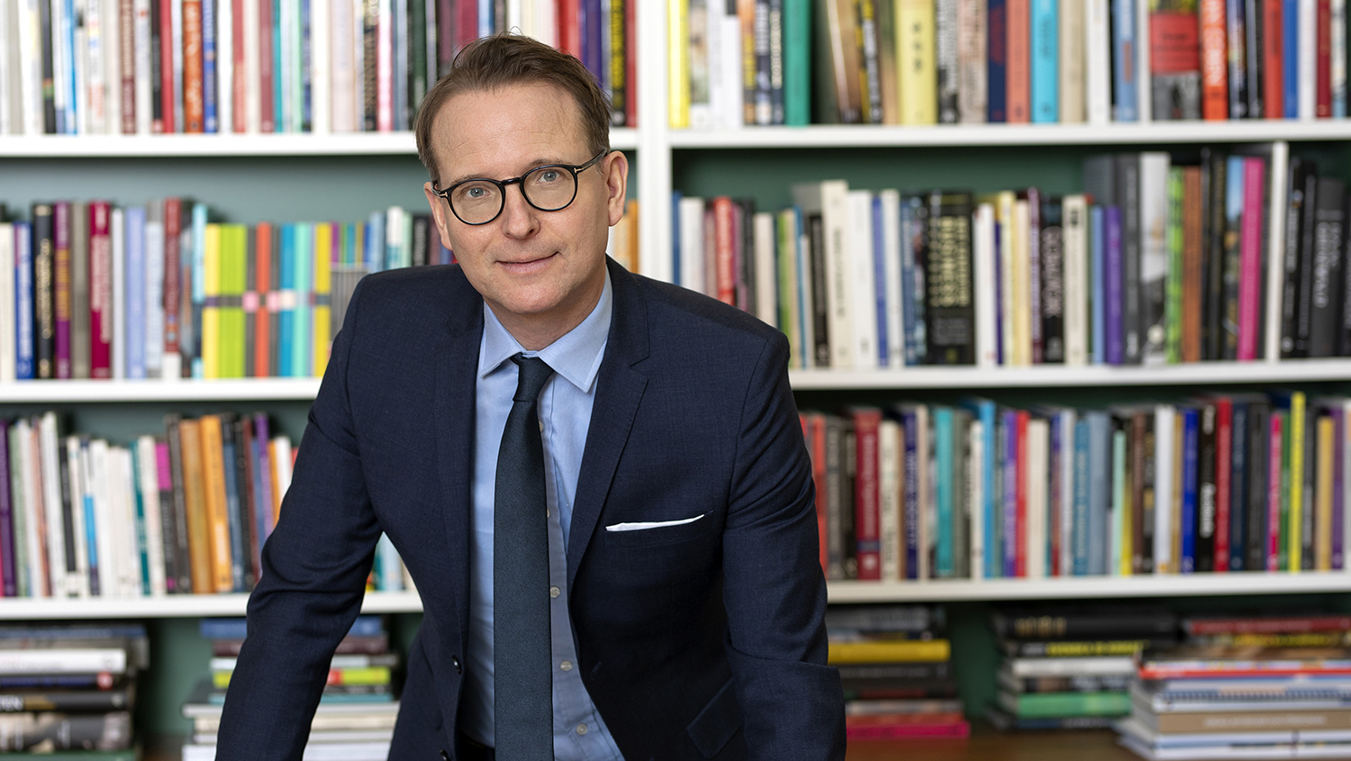 Lars Strannegård i kostym och slips står framför en bokhylla, lätt lutad över ett bord.